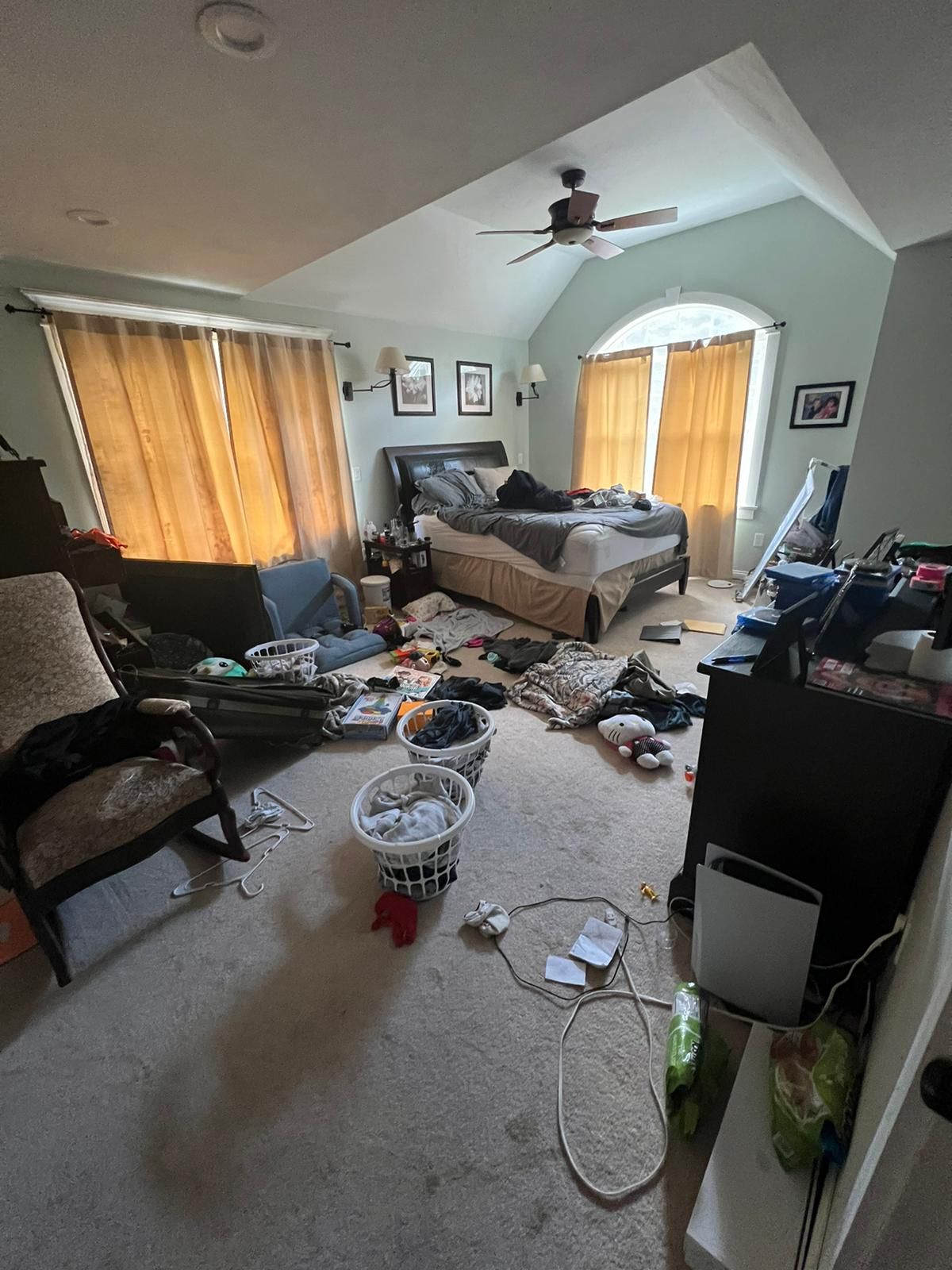 Cluttered bedroom with bed, furniture, and clothes strewn about. Beige walls, curtains, and carpet.
