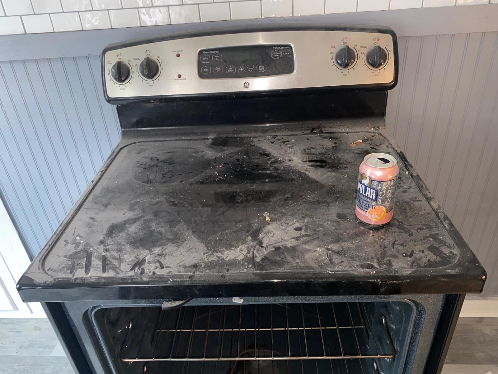 Dirty stovetop with a soda can, oven door open. Black and silver appliance in a kitchen setting.