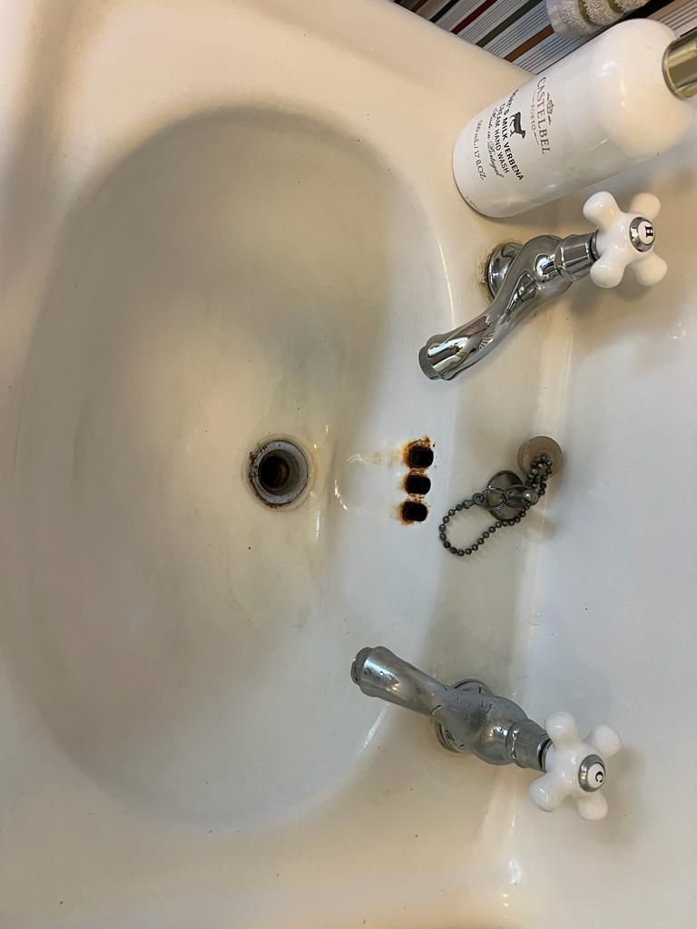 White bathroom sink with chrome faucets and a drain. A bottle rests on the rim.