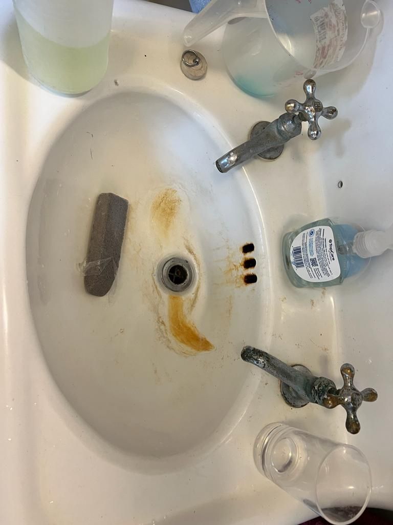 White, stained sink with two faucets, soap, and containers.