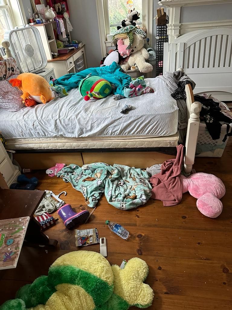 Messy bedroom with unmade bed, clothes, and stuffed animals strewn about.