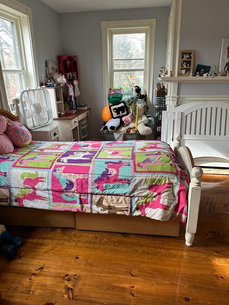Bedroom with white bed, colorful quilt, desk, and window with natural light.