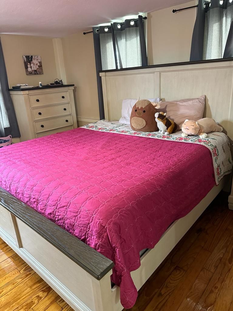 Bedroom with a large bed covered in a pink quilt and pillows. A chest of drawers is visible.
