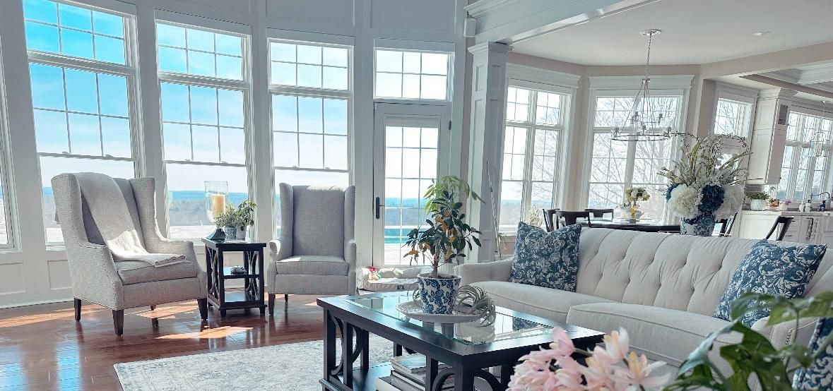 Living room with white sofa, armchairs, and many windows overlooking a bright blue sky.