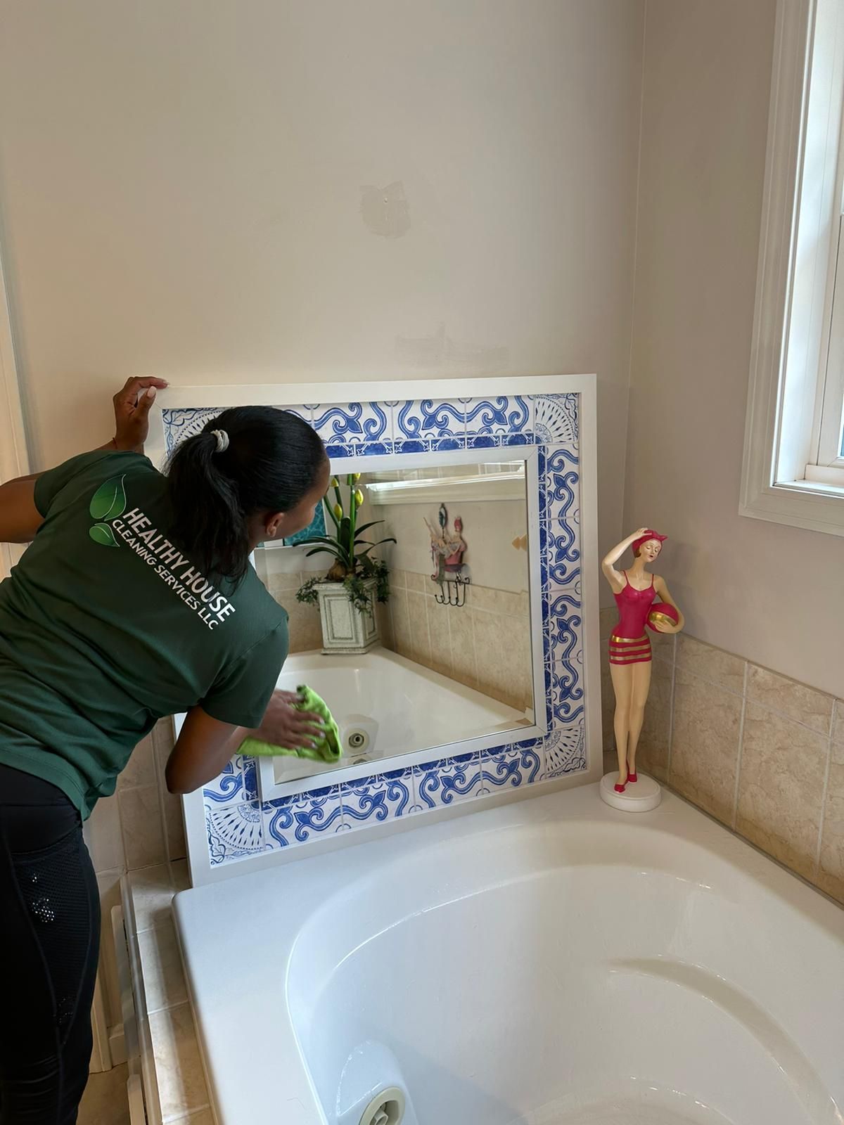 Person cleaning a bathroom mirror over a tub, wearing a green shirt, a decorative figurine is to the right.