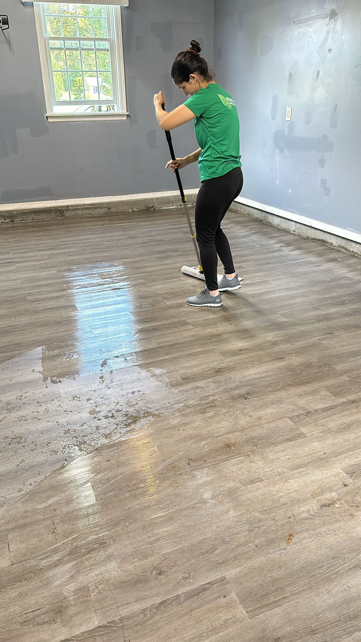 Person in green shirt mopping a wet floor in a room with a window.