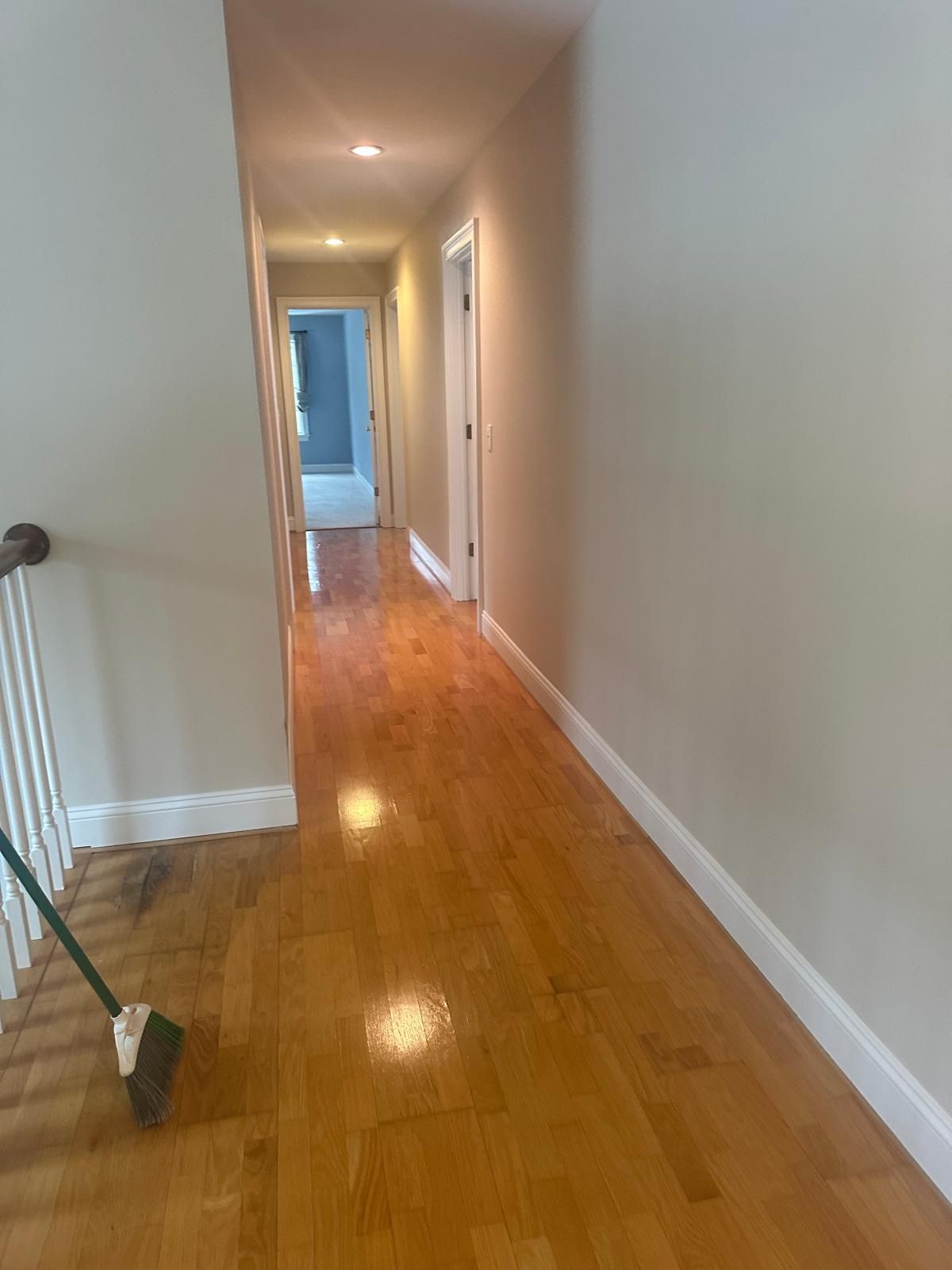 Hallway with hardwood floor and beige walls, broom on left. Doorways visible in the distance.