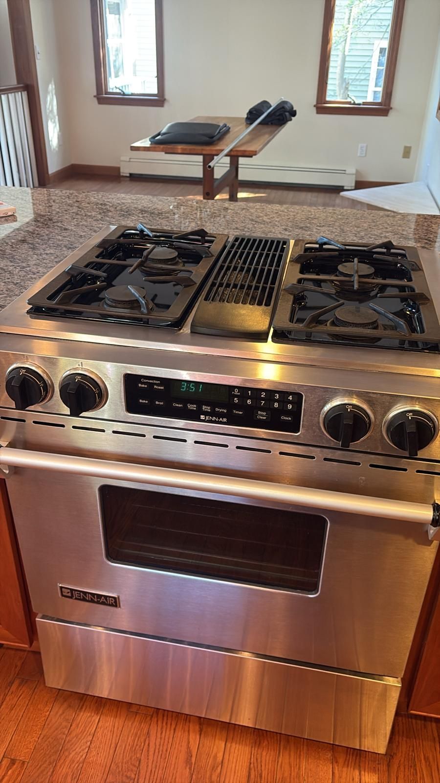 Stainless steel gas range in kitchen. Granite countertop, windows in background.