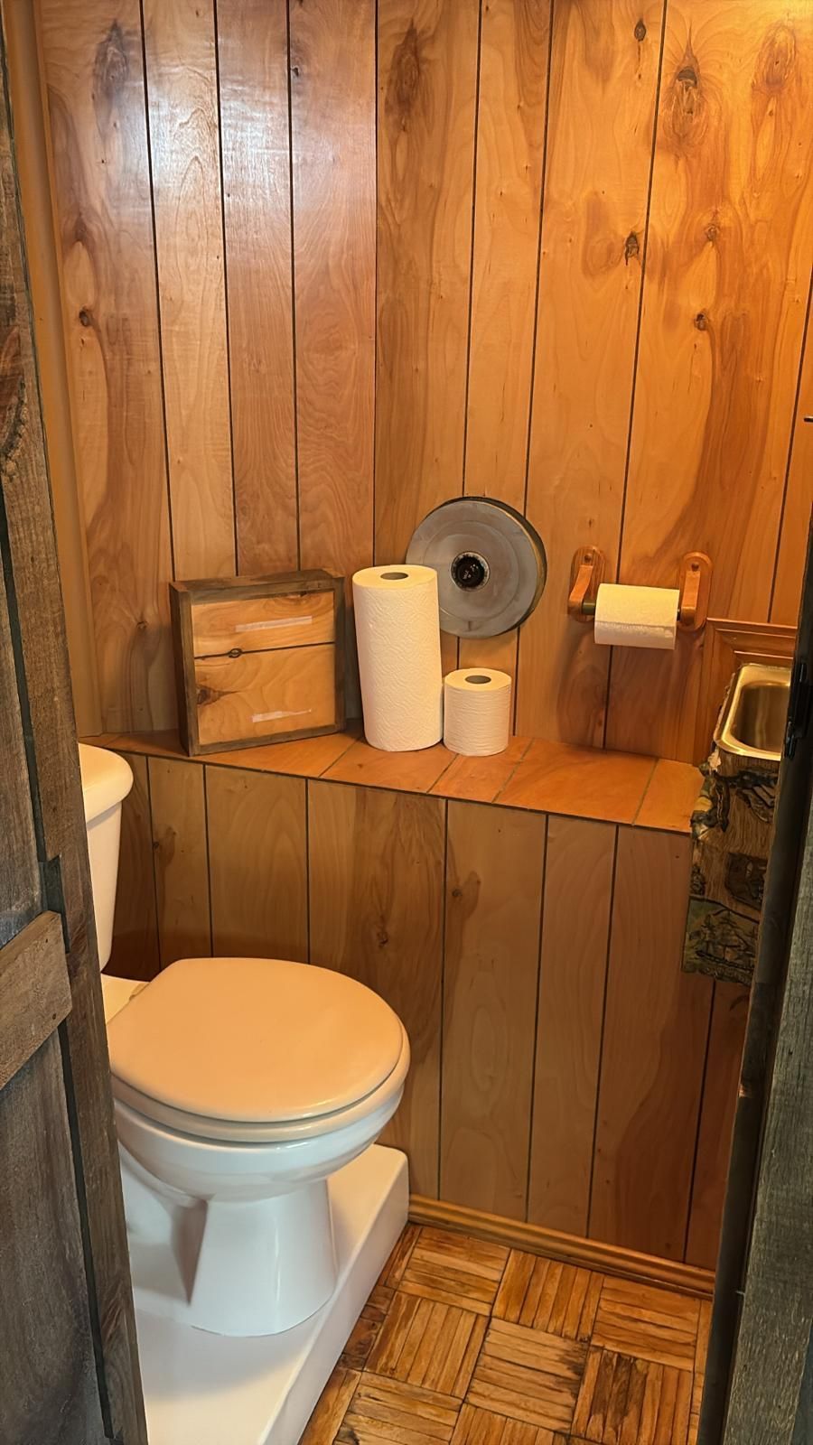 Small wooden-paneled bathroom with a toilet, shelf with supplies, and a paper towel holder.