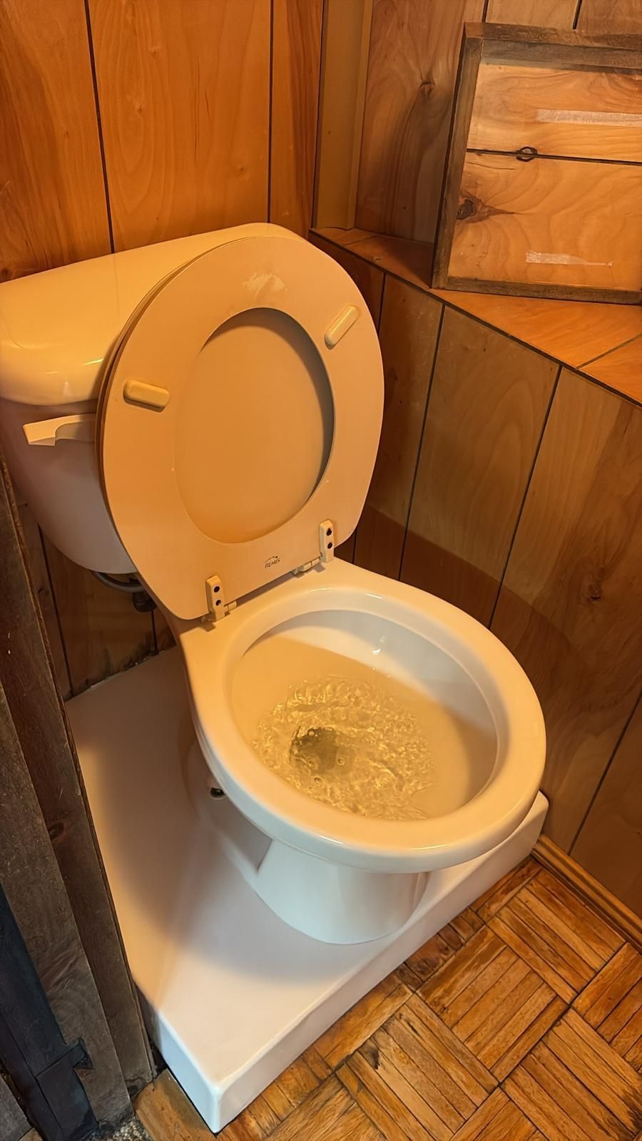 Toilet with open lid, water swirling inside. Set in wood-paneled bathroom, mounted on white base.