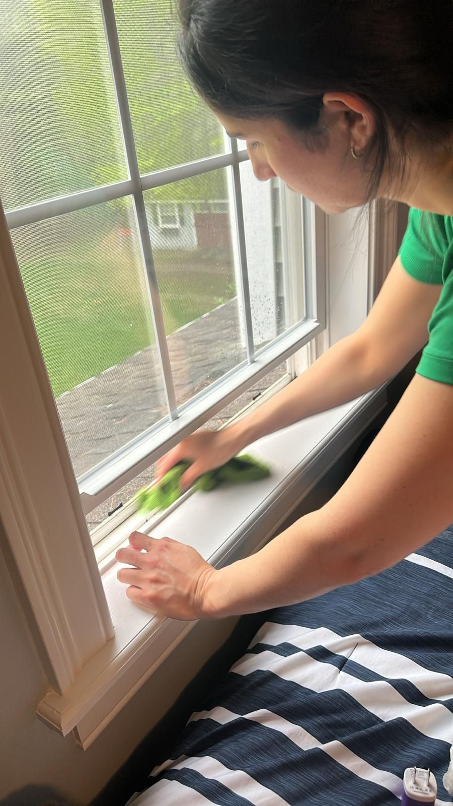 Person cleans a window sill with a green cloth. Window is white with a screen.