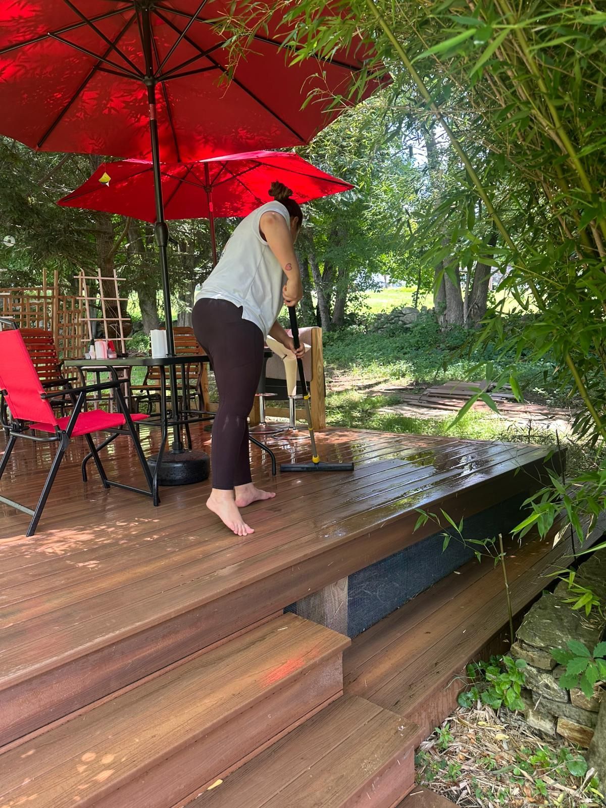 Person cleaning a wood deck under a red umbrella. There is outdoor furniture, greenery, and steps leading to the deck.