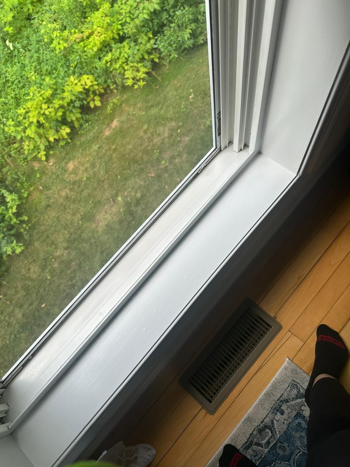 A window looking out at green trees and grass. A white windowsill with a vent sits above wood flooring.