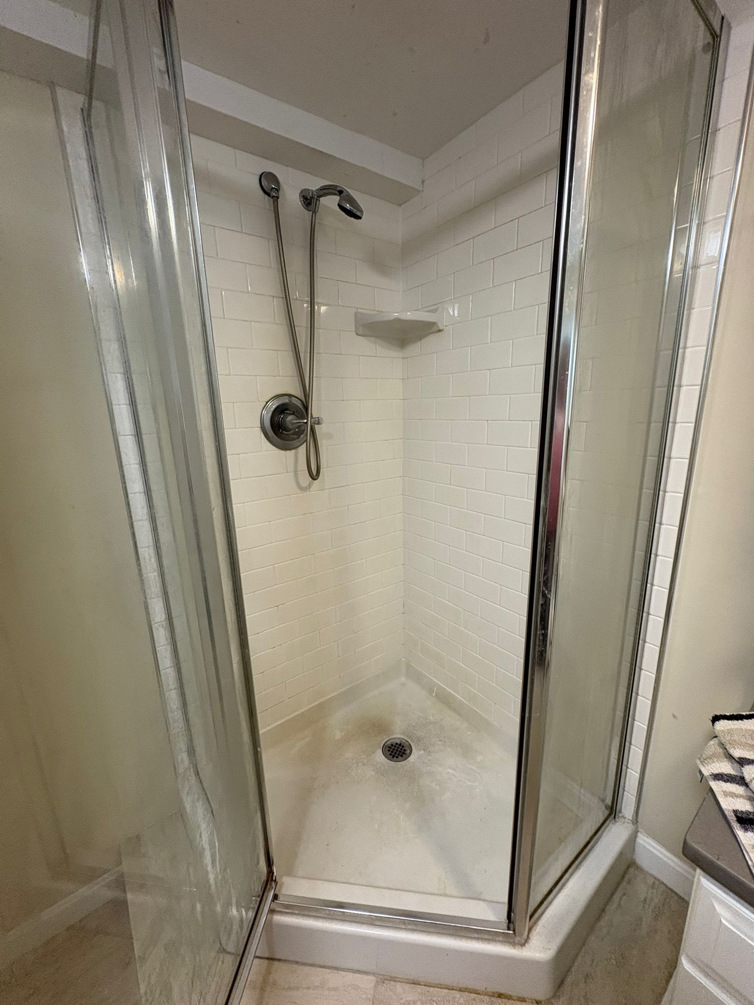 A corner shower with white tiled walls and glass doors. Visible showerhead, soap shelf, and some stains.