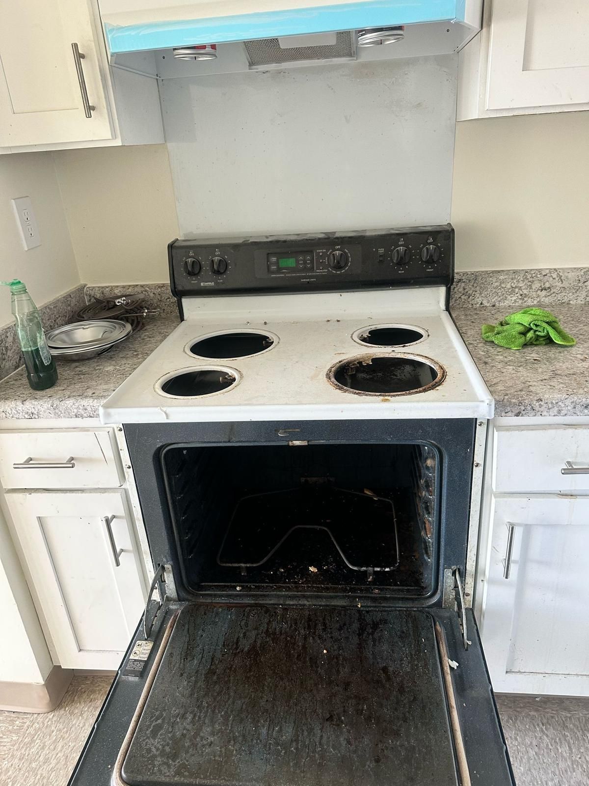 An open, dirty oven with a black stovetop, surrounded by white cabinets.