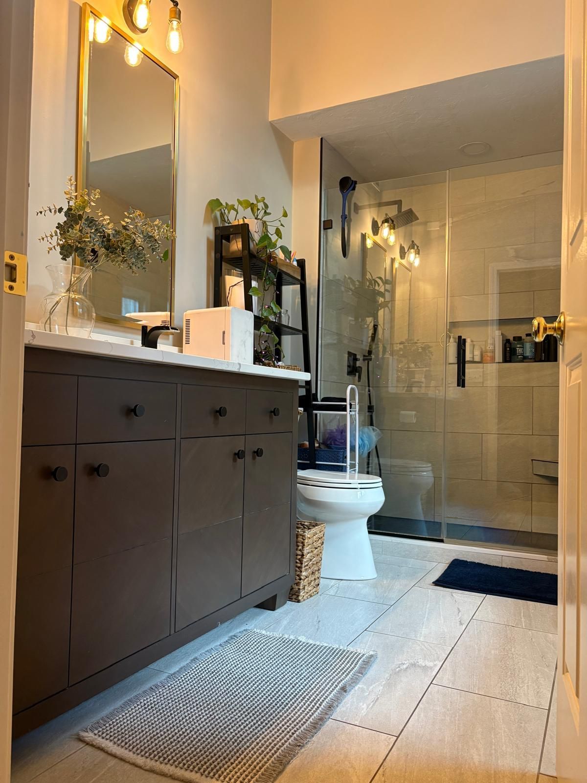 Bathroom with dark brown vanity, large mirror, glass shower, and neutral tile flooring.