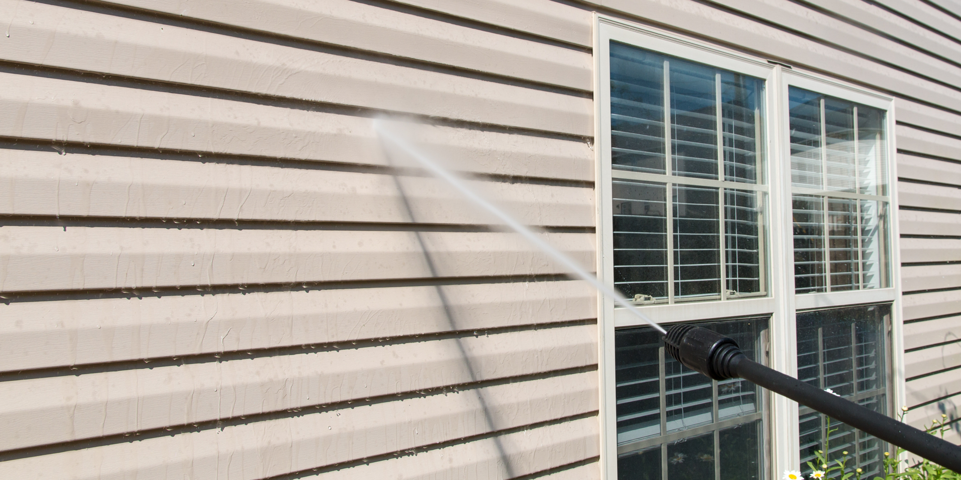 A person is power washing the side of a house with a window. The siding is being cleaned.