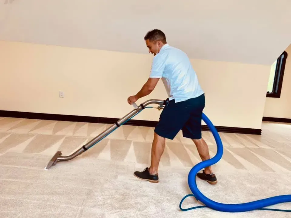 Man cleaning a carpet with a machine in a room with beige walls and a light-colored carpet.