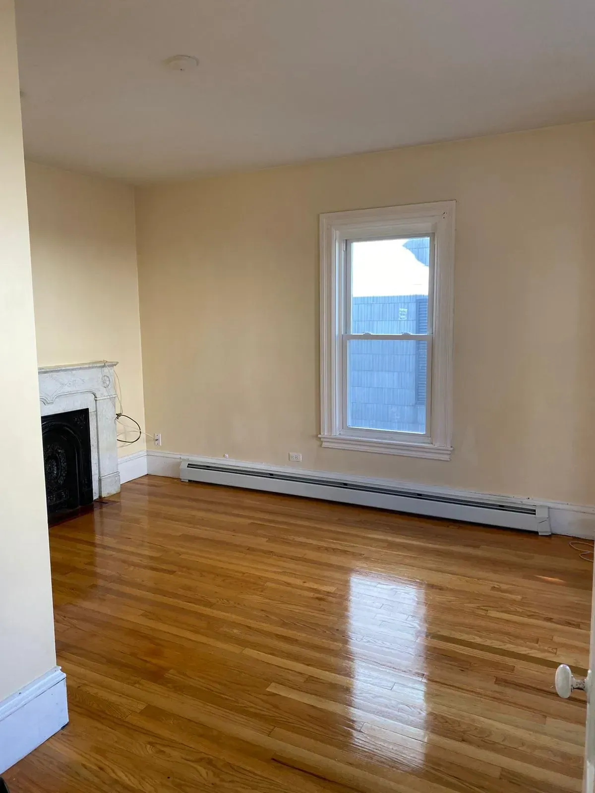 Empty room with hardwood floors, window, and a fireplace. Walls are beige.