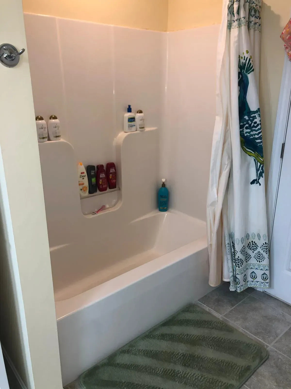 White bathtub and shower surround with toiletries, green rug, and patterned shower curtain.