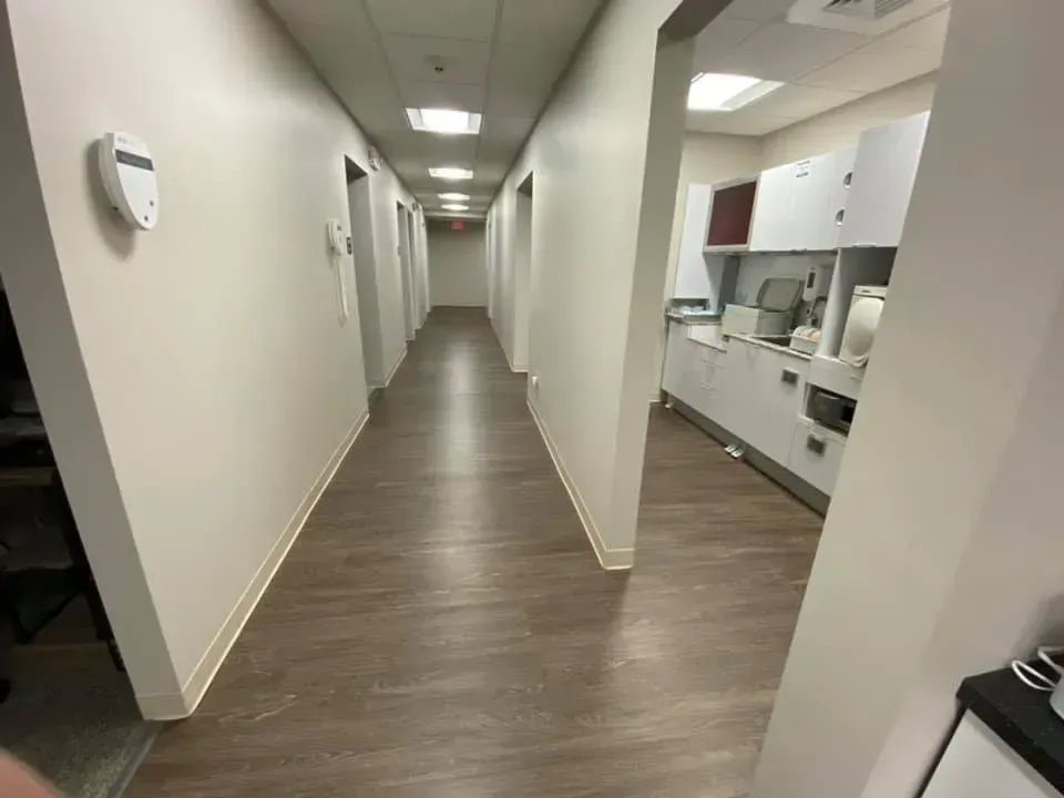 Long, empty hallway with white walls, wood-look flooring, and recessed lighting. A doorway leads to a bright lab space.