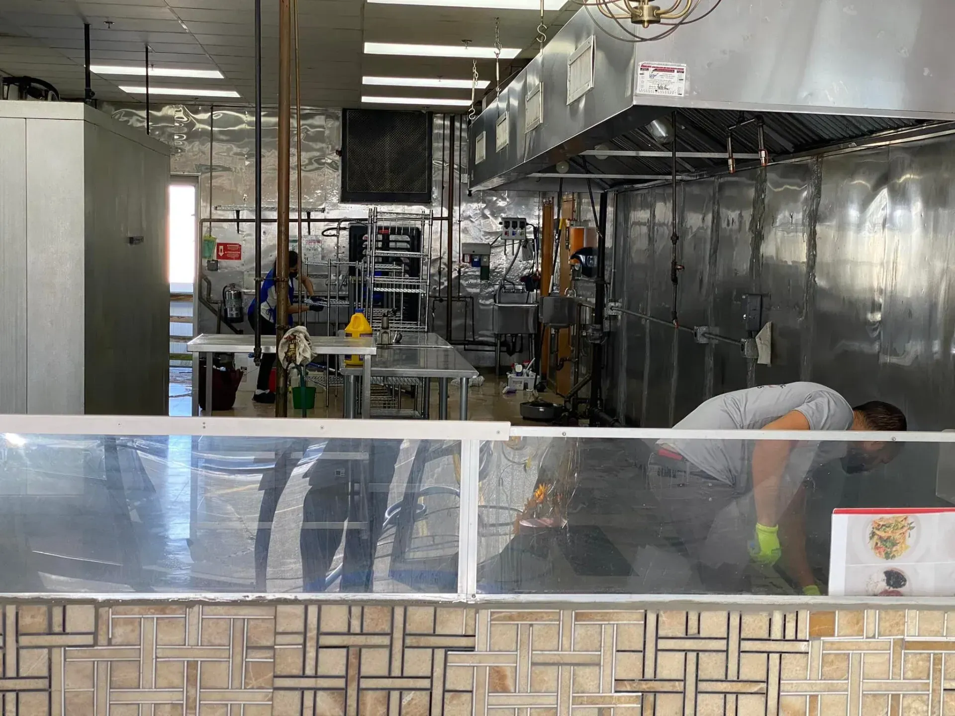 Kitchen interior with workers; stainless steel appliances, ventilation hood. Someone cleaning the floor.