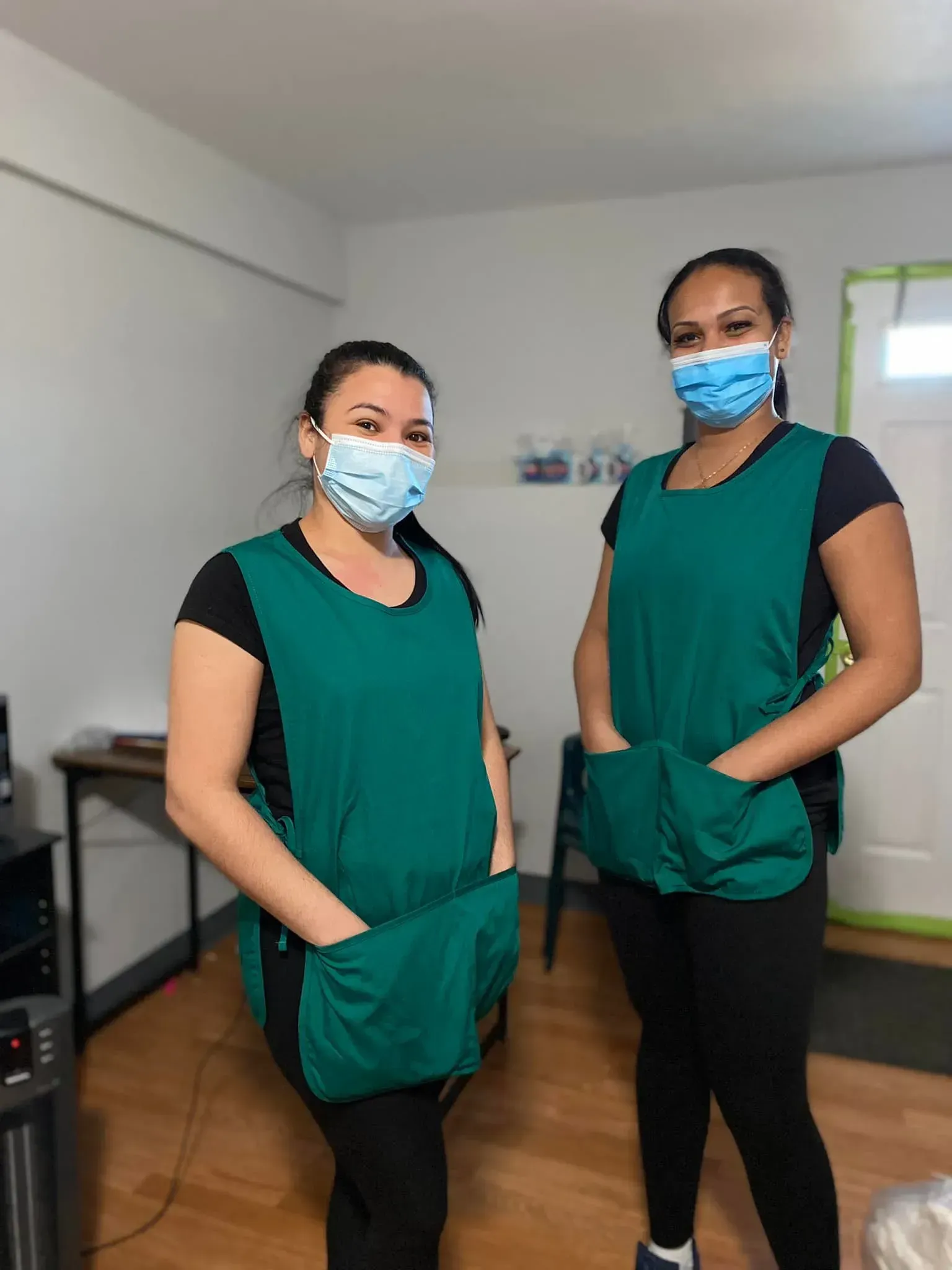 Two women wearing teal aprons and masks, standing indoors with hands in pockets.