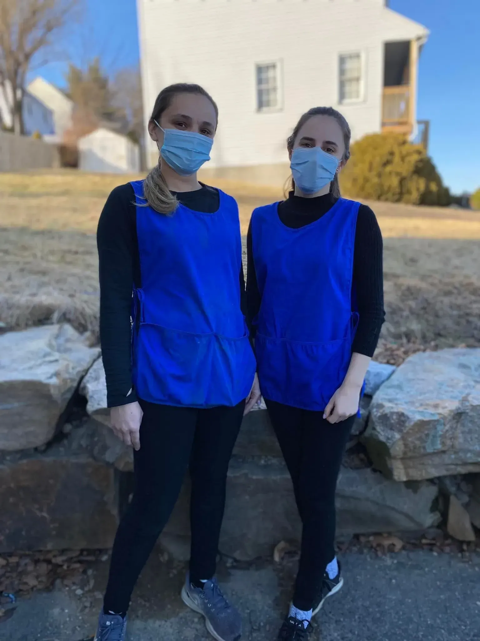 Two people in blue vests and masks stand outside a building.