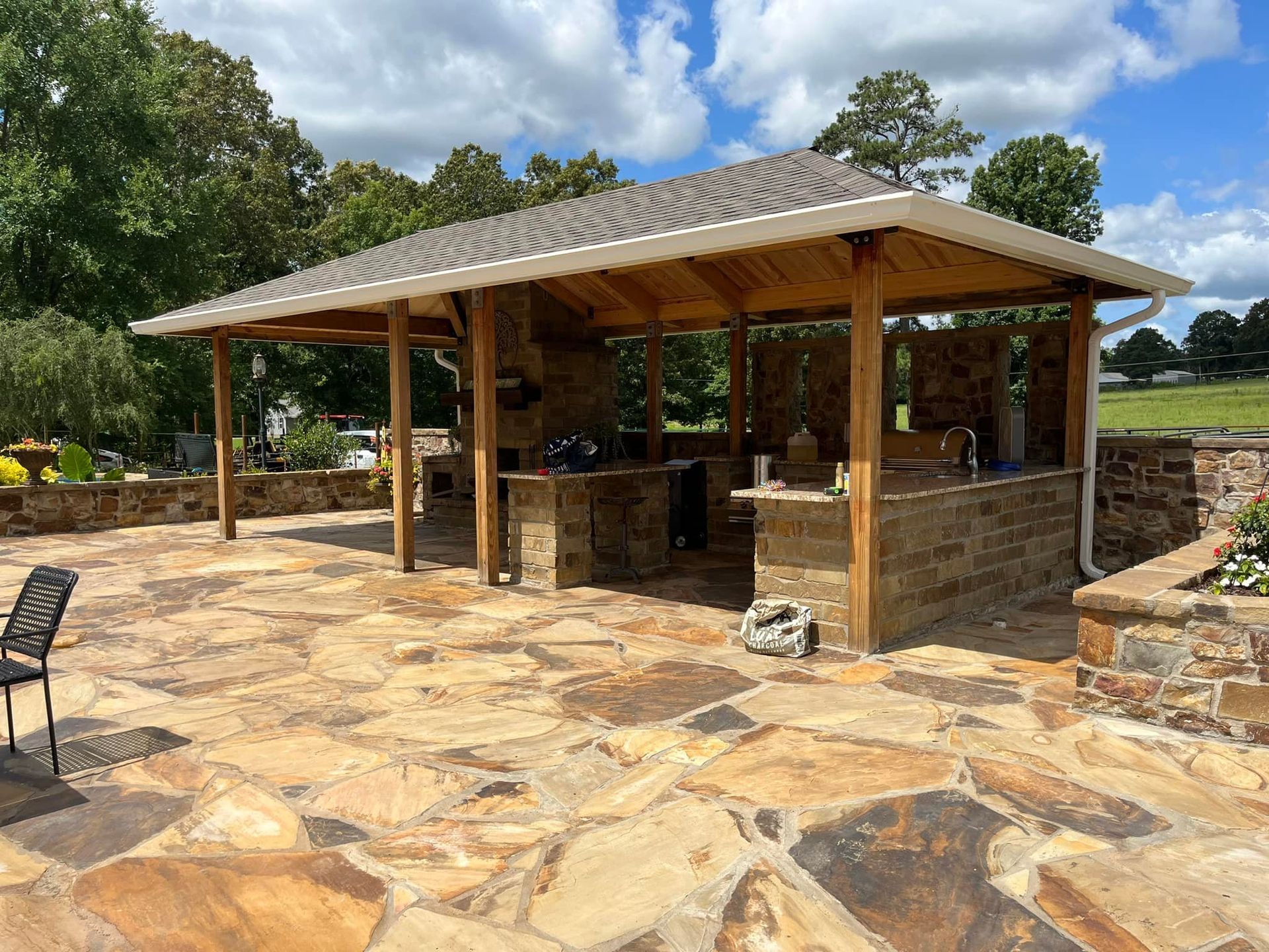 A large outdoor kitchen with a wooden roof is sitting on top of a stone patio.