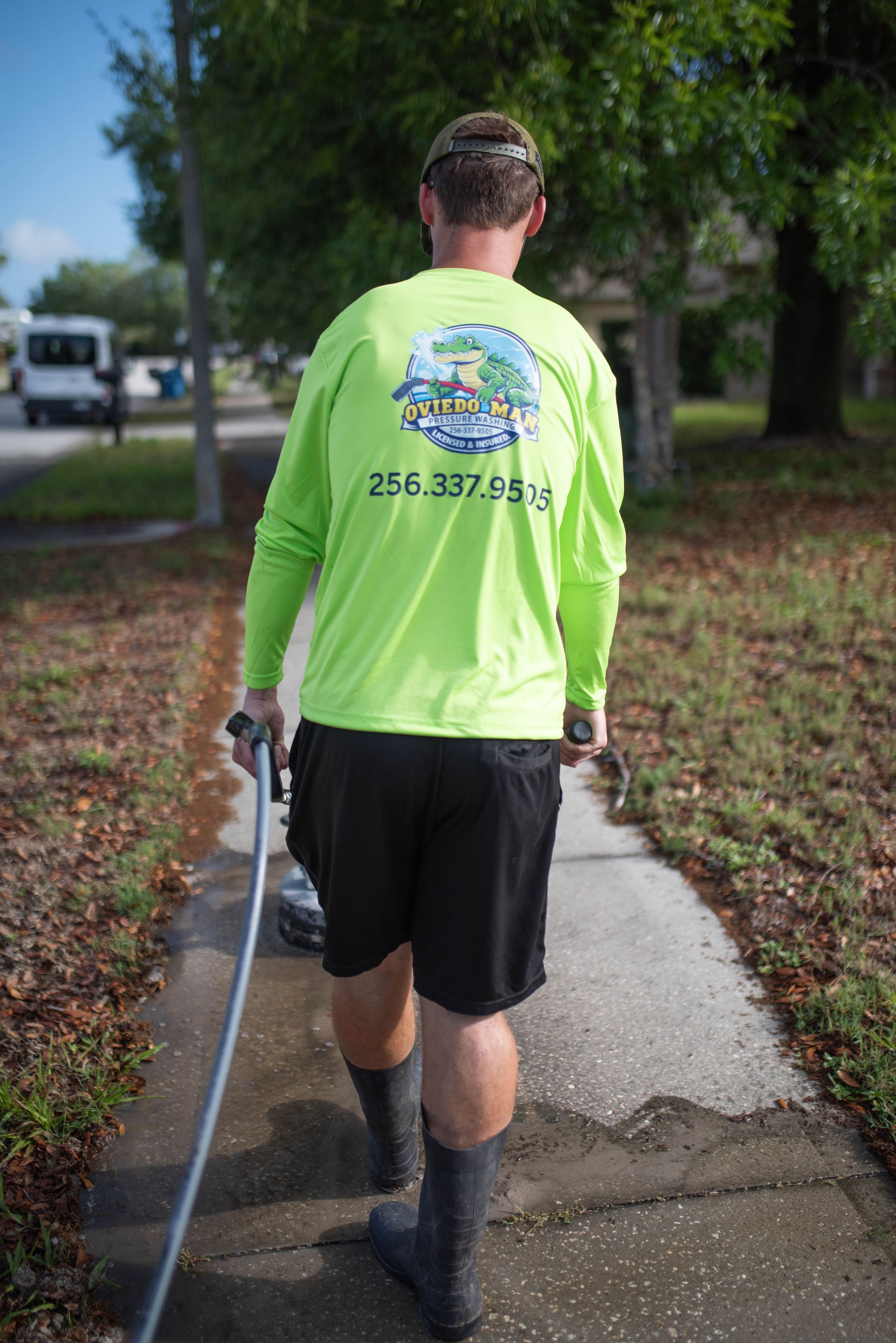 A worker in a neon green long-sleeve shirt walks along a sidewalk, holding a hose while performing outdoor maintenance.