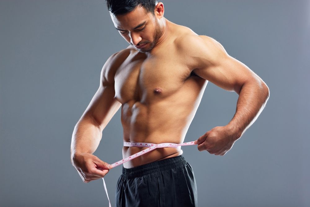 A shirtless man is measuring his waist with a tape measure.