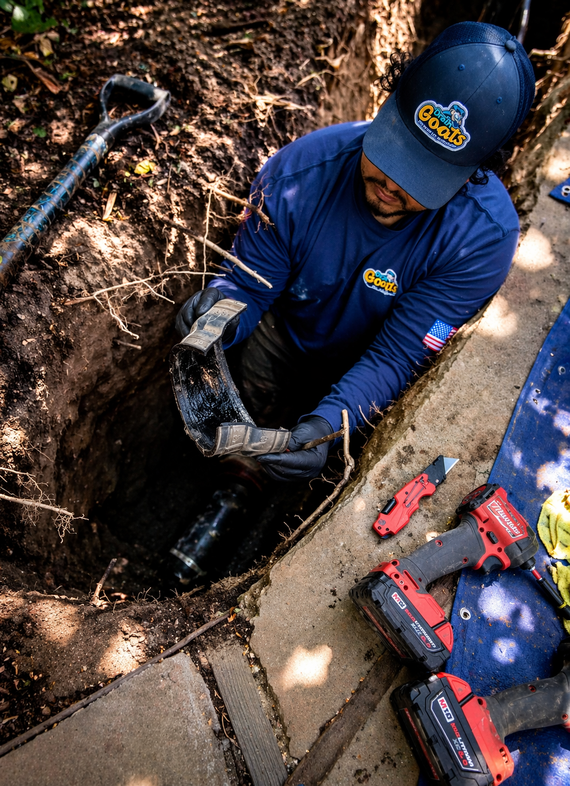 Plumbing pipes in a dirt hole, connected with fittings. White PVC pipe and tools on the edge of the hole.