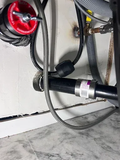 Plumbing under a sink: black pipe, stainless steel hose, red device with a knob, and other lines.