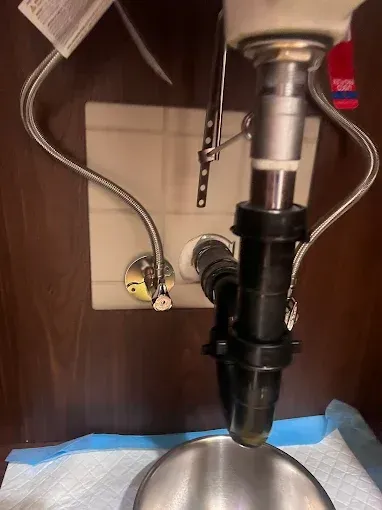 Plumbing under a sink: chrome faucet connections, black drainpipe, and a stainless steel bowl sitting below.