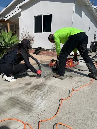 Two people cutting and vacuuming concrete on a driveway; one using a saw, the other a vacuum.