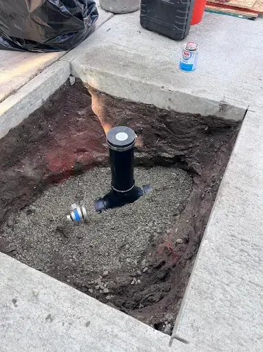 Hole in concrete with sprinkler head, gravel, pipes, and tools.