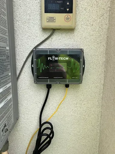 Flow-Tech water conditioner on a wall, showing a green waveform display. Two cables, black and yellow, connected below.