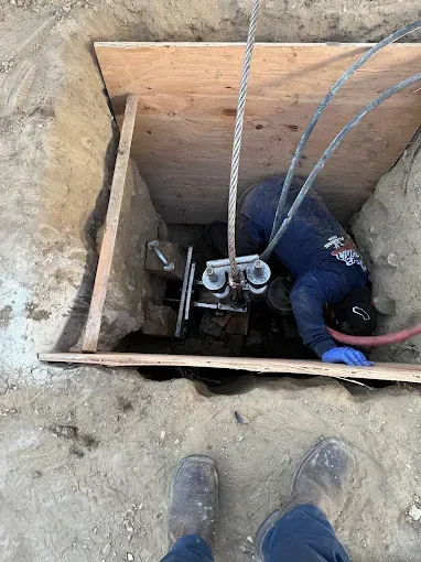 Person working in a deep trench, using equipment. Protective wooden frame and gray cables overhead.