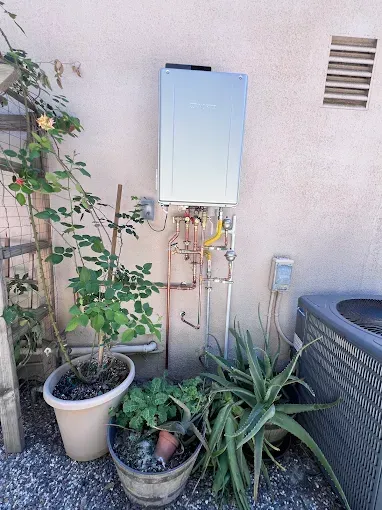 Outdoor water heater on a stucco wall, with copper pipes, and potted plants in front.