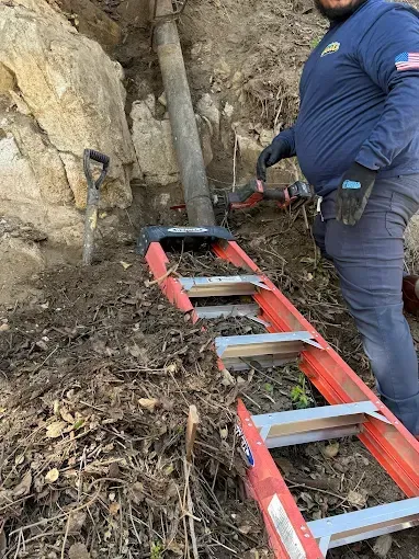 Man working, using a ladder and tools, alongside a large pipe embedded in dirt and rock.