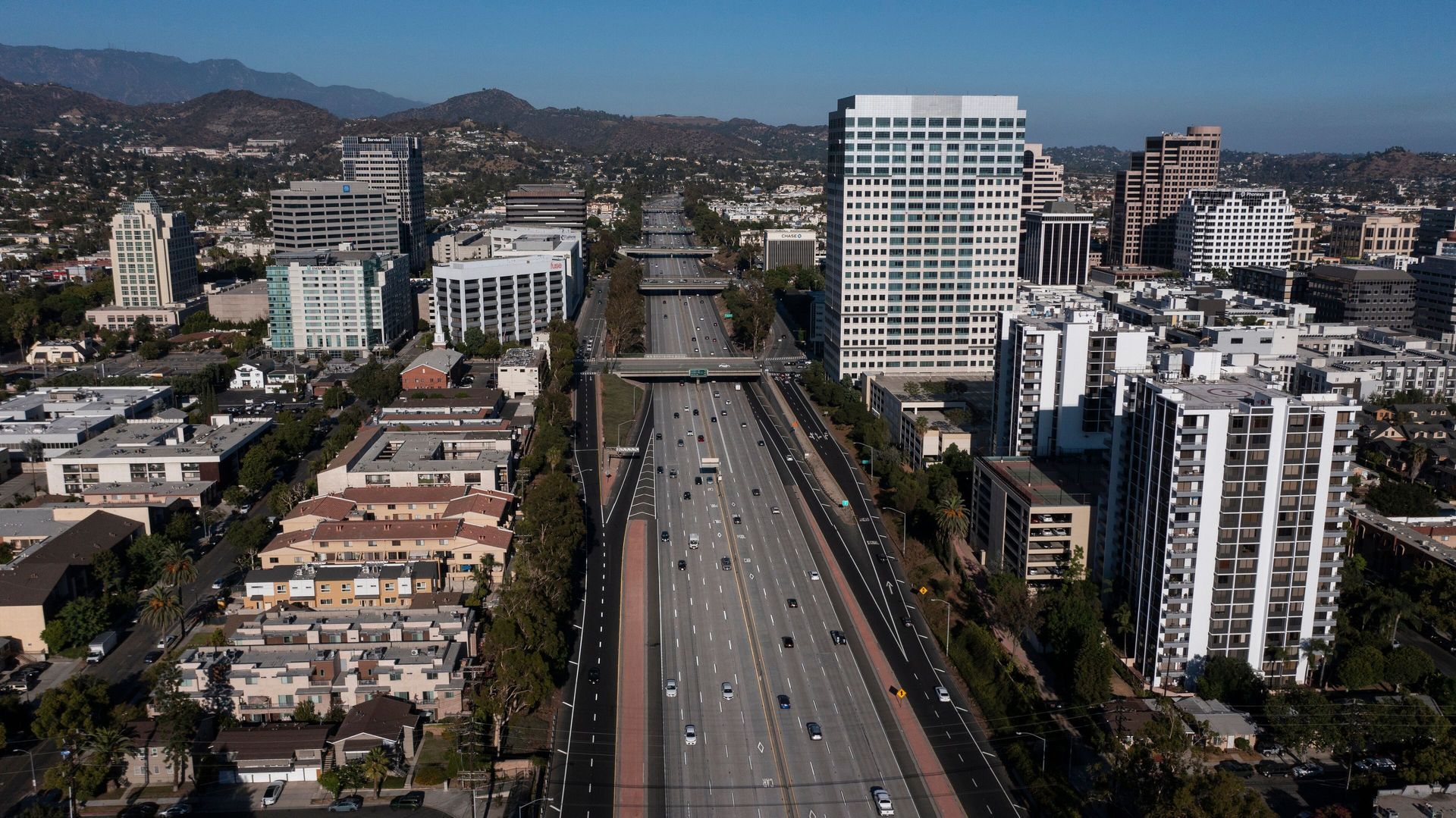Aerial view of a Los Angeles highway with cars, surrounded by buildings and a mountain range under a clear blue sky.