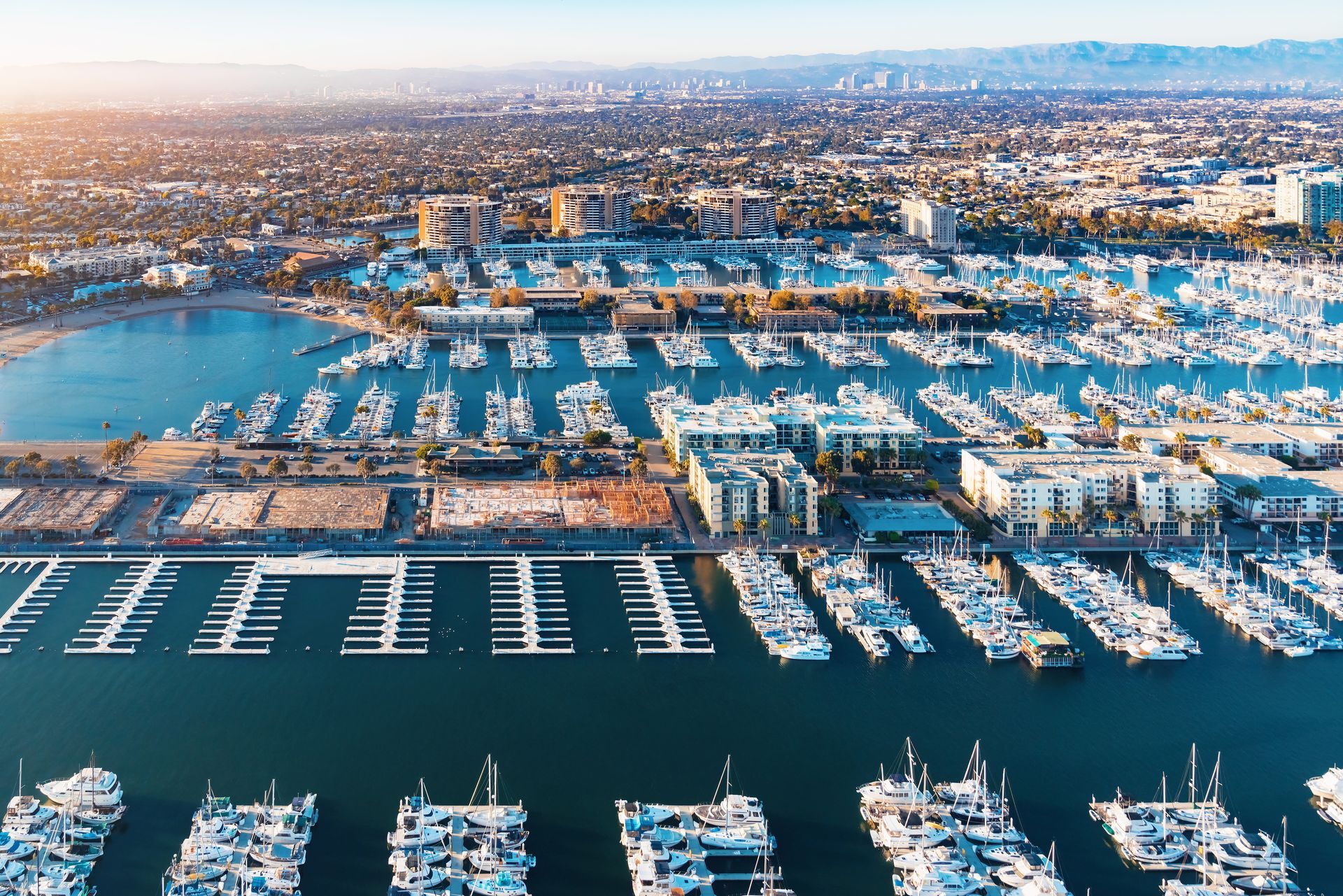 Aerial view of a marina filled with boats. Buildings and city skyline are visible in the background under a sunny sky.