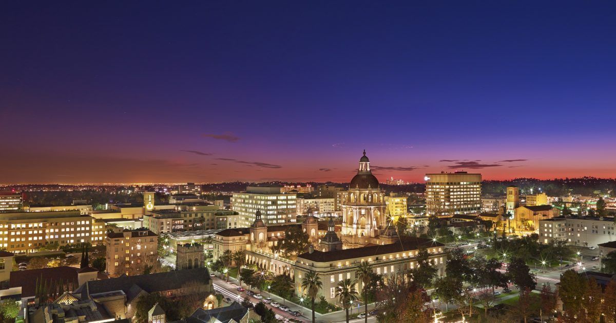 Nighttime cityscape with buildings illuminated against a twilight sky, trees in the foreground.