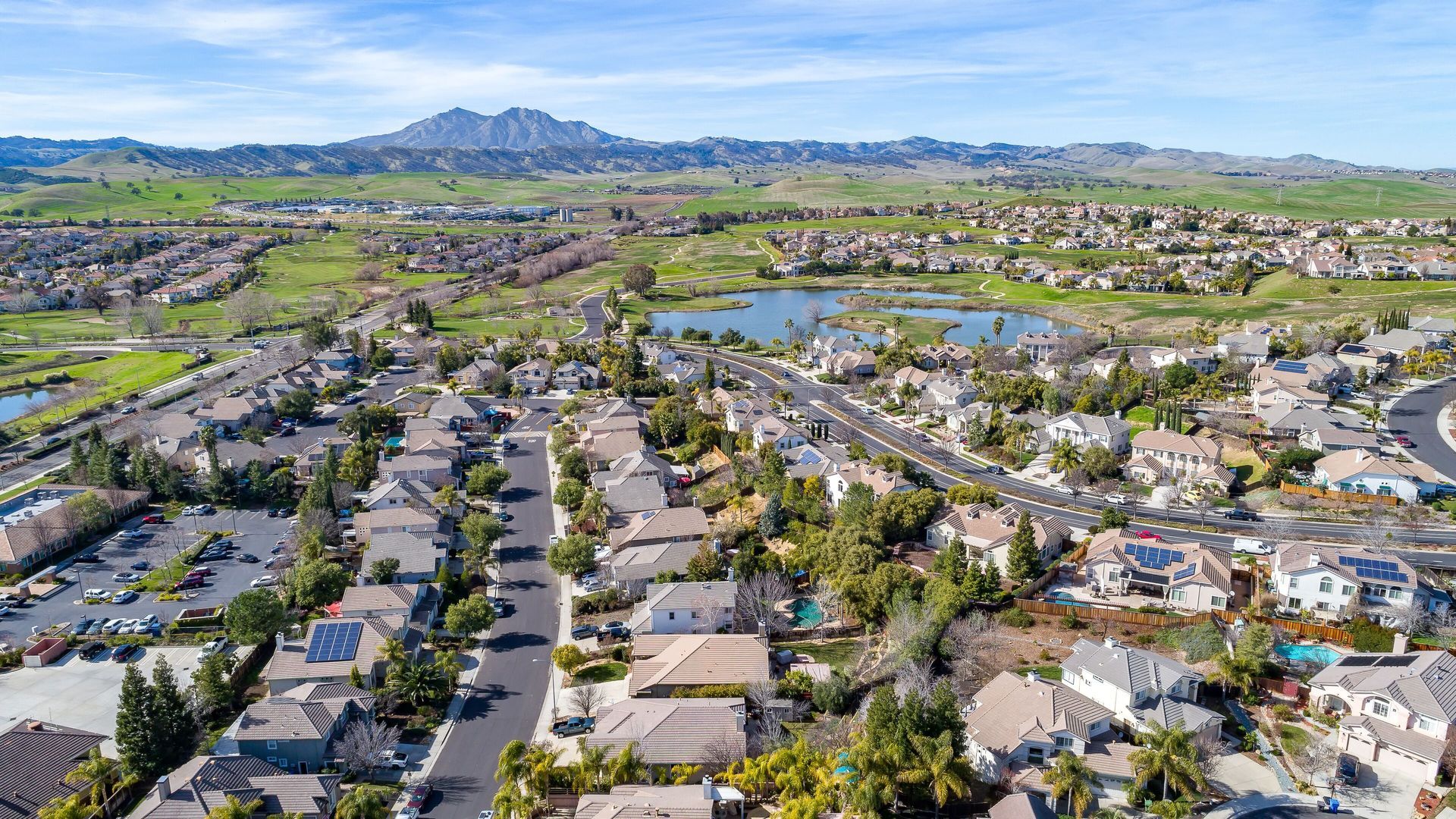 Aerial view of suburban homes, roads, a lake, golf course, and mountains under a blue sky.
