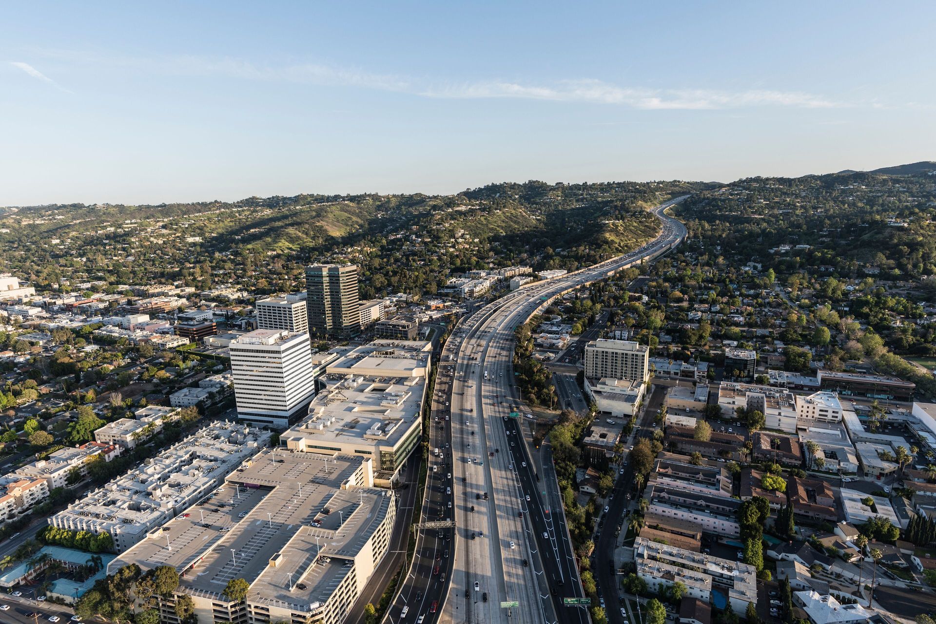 Aerial view of a Los Angeles highway cutting through a cityscape and hilly, green terrain under a clear sky.