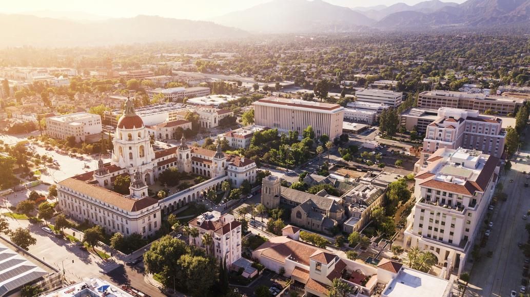 Aerial view of a city with ornate buildings, trees, and mountains in the background, bathed in sunlight.