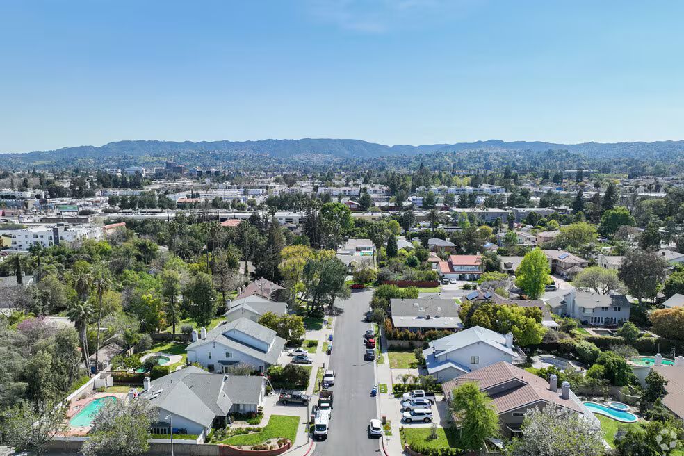 Aerial view of suburban neighborhood with houses, trees, and street, mountains in the distance under a blue sky.