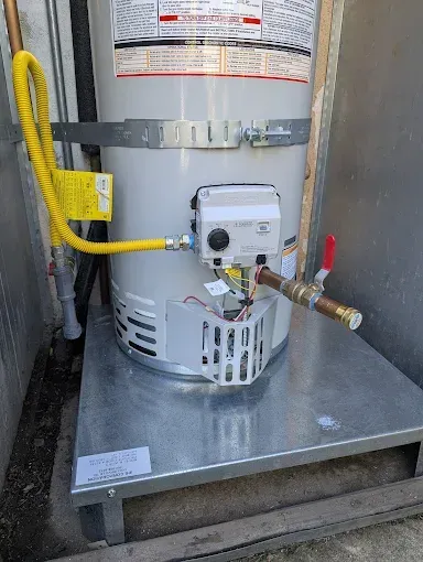A gas water heater on a metal platform, outdoors. Yellow gas line connects to the unit's valve.