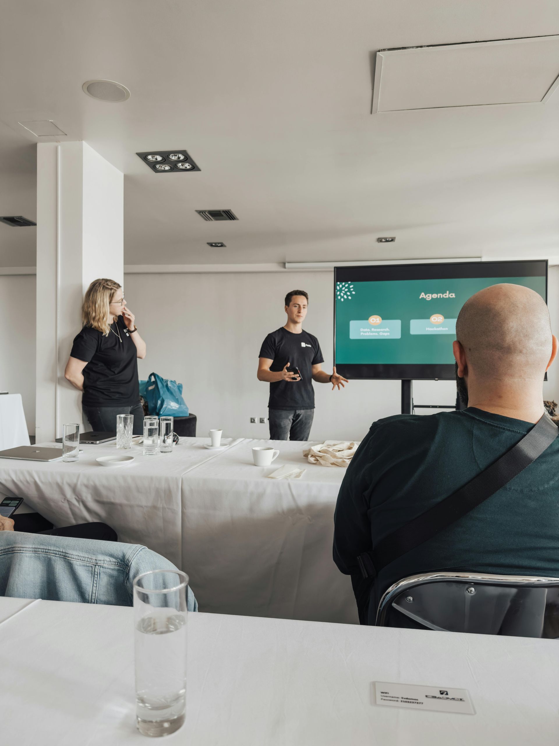 Speaker presenting in a meeting room with a slide on a screen, audience seated at tables