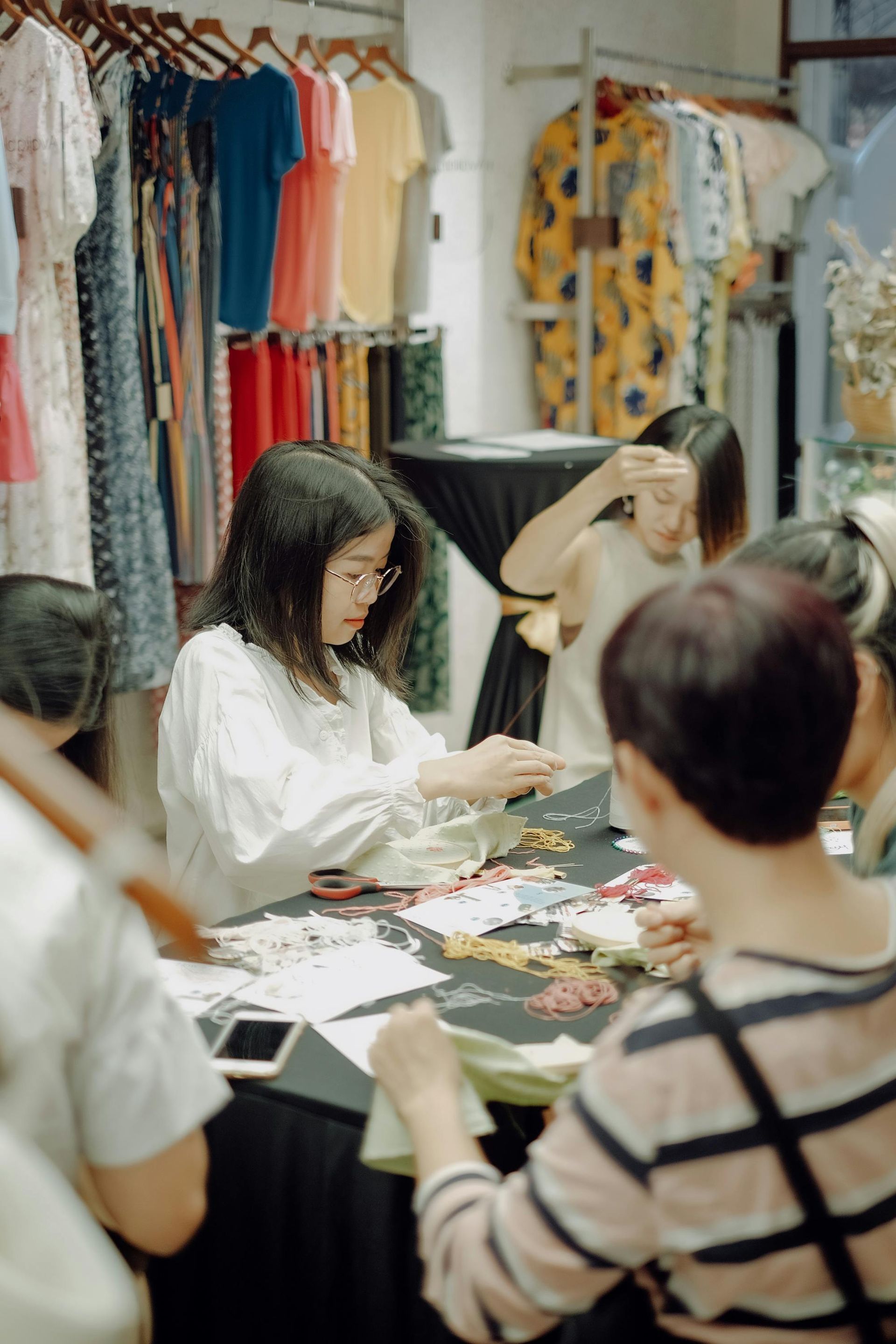 People seated around a table, crafting or sorting items in a clothing shop with racks of colorful garments.